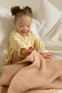 A smiling toddler in a yellow long-sleeved shirt sits on a bed, reaching for a tan, waffle-textured superfine merino The Popcorn - Henna baby blanket.