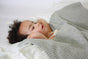A child with curly hair lies on a white pillow, covered by a gray, textured The Popcorn Blanket - Moon Stone.