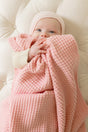 A baby in a light pink bonnet is wrapped in a soft, textured Pink Quartz Popcorn Blanket by AU Baby.