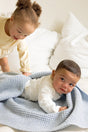 A toddler reaches toward a baby lying on a light blue, textured AU Baby The Popcorn Blanket - Sky Blue.