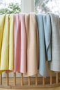 Five folded, plant-dyed merino baby blankets, including The Sawa - Pink Quartz, in yellow, pink, peach, blue, and gray draped over a wooden rack.