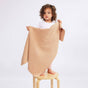 A toddler stands on a wooden stool holding up The Sawa - Henna, a textured, light brown merino baby blanket with personalized embroidery.