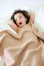 A child with their hands behind their head rests under a tan, plant-dyed AU Baby The Sawa - Henna merino baby blanket on a white bed.
