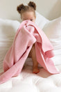 A toddler peeks from behind a draped, plant-dyed pink merino baby blanket, The Sawa - Pink Quartz by AU Baby, while sitting on a white tufted surface.
