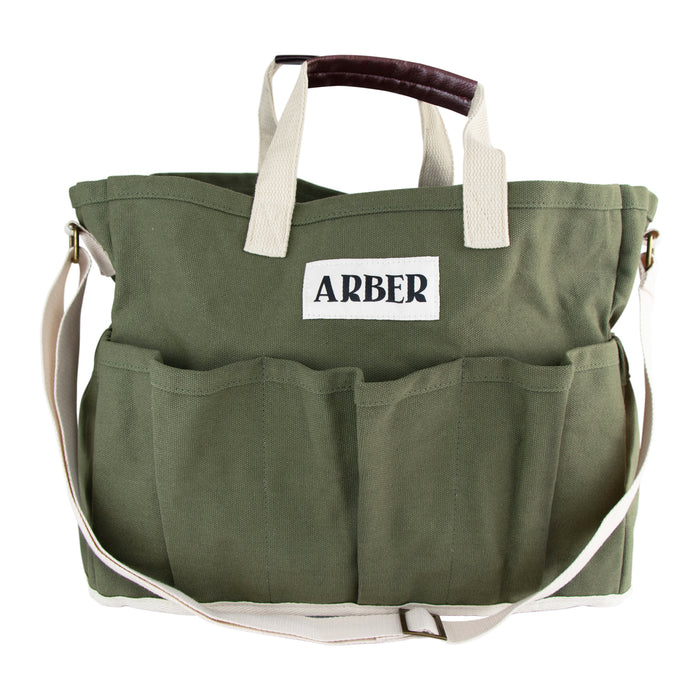 The Arber Garden Tote is a green canvas gardening bag featuring a spacious compartment, six exterior pockets, an adjustable shoulder strap, and short beige handles with a dark brown grip. It is adorned with a white ARBER label for style and comfort.