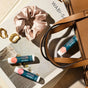 Three Cliganic Tinted Lip Balms (Hint of Tint), a beige scrunchie, gold earrings, and a tan handbag rest atop a Wabi-Sabi book, all arranged on a neutral surface in sunlight.