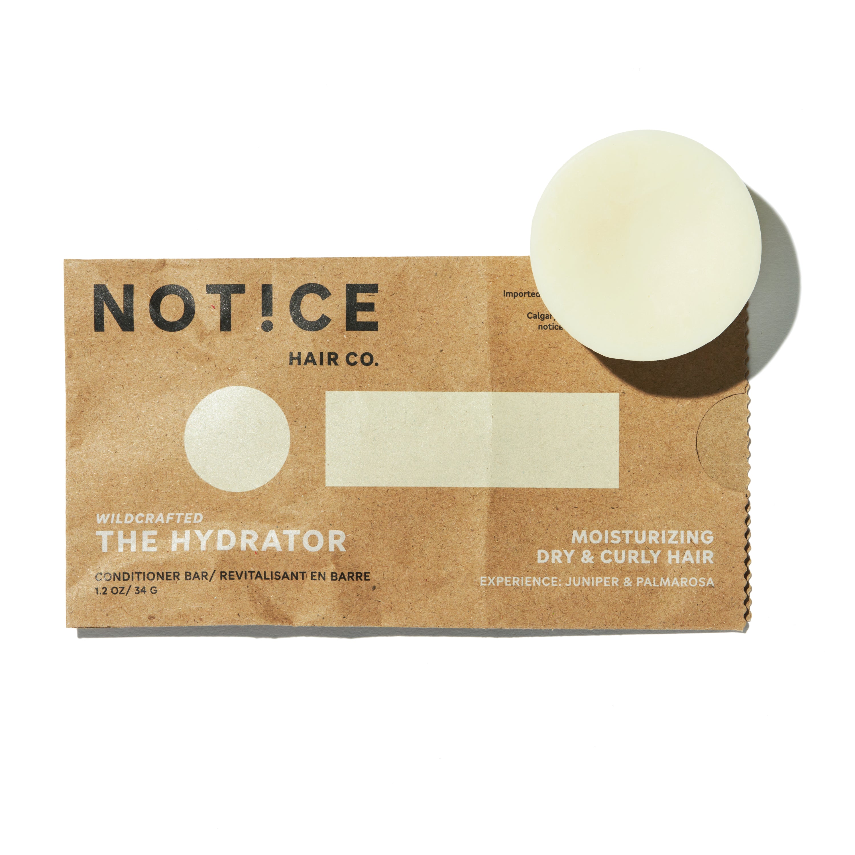 A round conditioner bar rests on an open brown paper wrapper labeled NOT!CE Hair Co. The Hydrator. This plastic-free hair care product hydrates dry and curly hair effectively with juniper and palmarosa, emphasizing its moisturizing benefits.