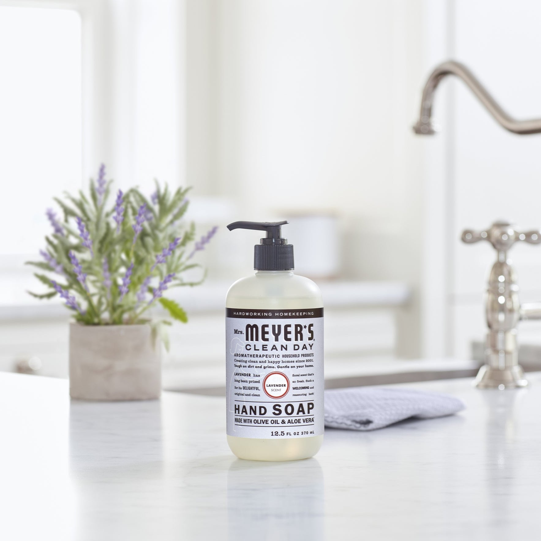 A Mrs. Meyers Hand Soap bottle, noted for its garden-inspired scents and essential oils, is on a white kitchen countertop. Behind it are a stainless steel faucet and a small lavender pot, with a gray cloth alongside the soap.