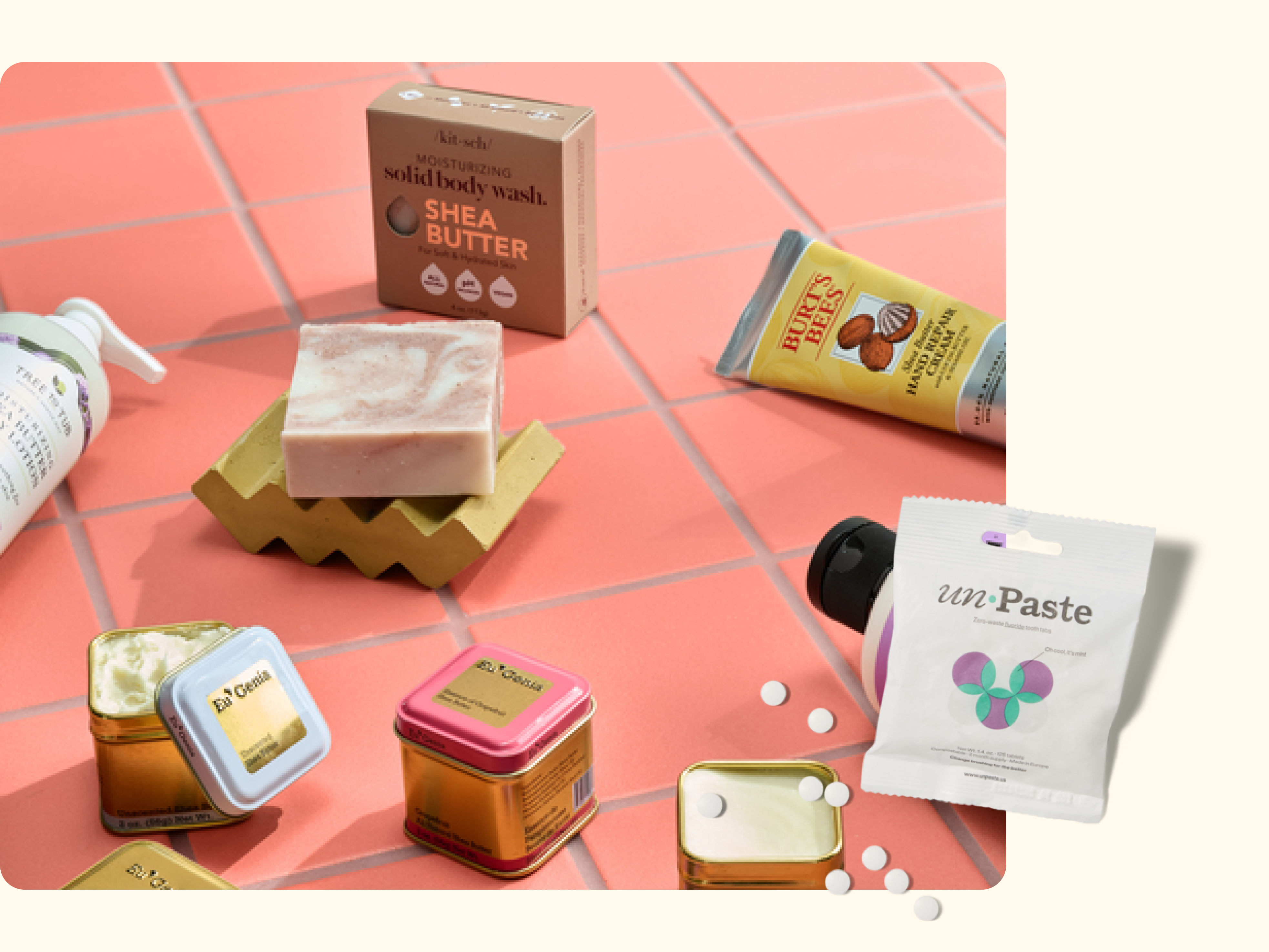 A collection of eco-friendly toiletries including a bar of shea butter soap, tins of deodorant, hand cream, toothpaste tablets, and a pump bottle, arranged on a pink tiled surface.