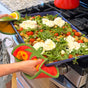 Wearing a gray apron and The Neighborgoods’ Pot Holder - Tomato, a person holds a baking dish of gnocchi, cherry tomatoes, greens, and cheese in front of the stove—a kitchen essential scene.