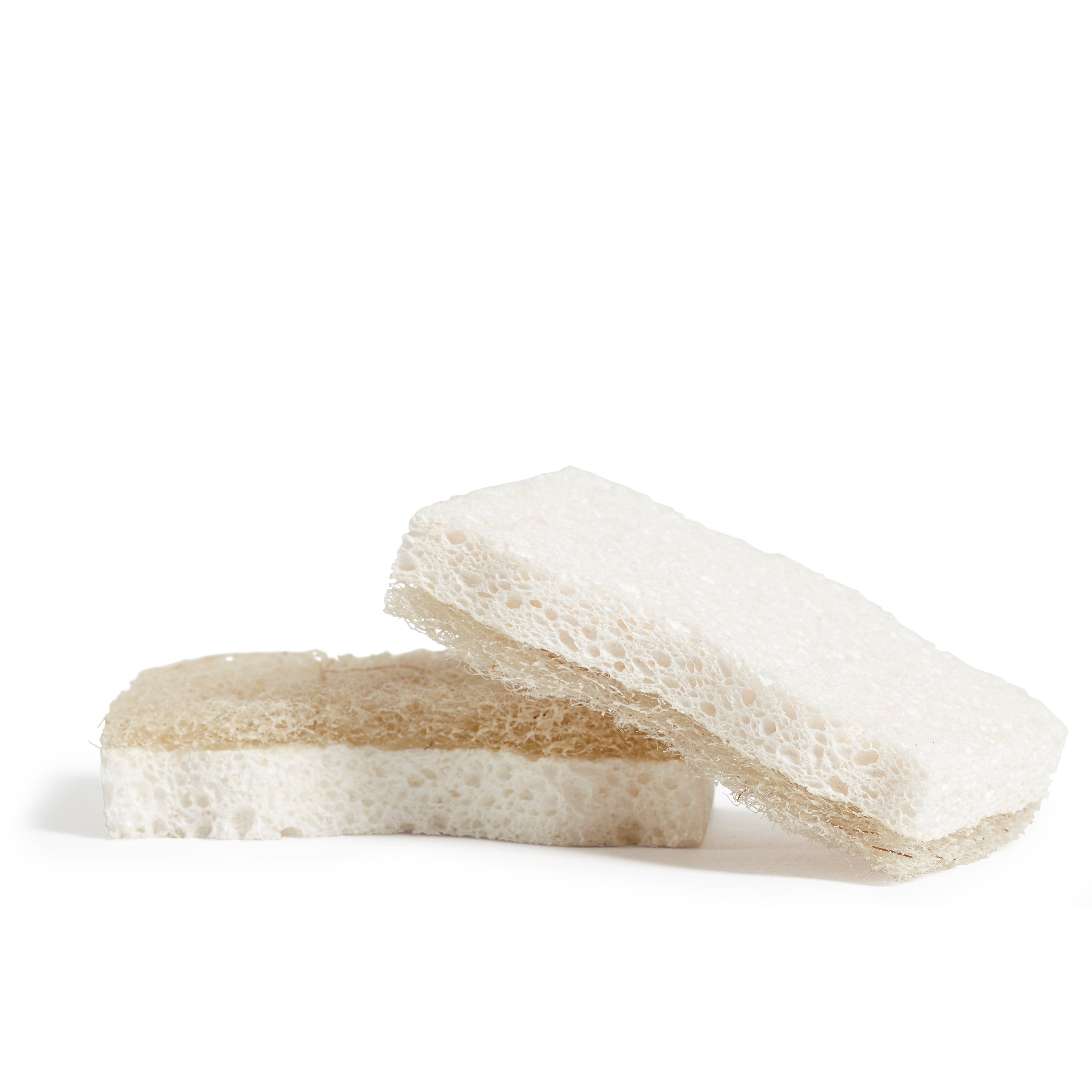 Two small, non-abrasive Coconut Scrubber Sponges by Grove Co. are stacked on a white surface. The top sponge is angled slightly, showcasing its textured, porous design; both sponges resemble coconut scrubbers and are light brown on one side and white on the other.