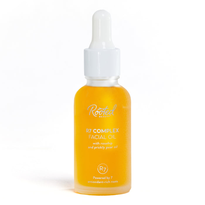 A clear glass dropper bottle filled with golden yellow facial oil, labeled Rooted Beauty R7 Complex Facial Oil with rosehip and prickly pear oil, standing upright on a white background.