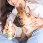 A woman in a white shirt sits holding a French Bulldog and a bottle of dental spray for pets, smiling as she prepares to use the product on the dog.