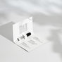 A small perfume sample vial attached inside an open white card displaying product information and scent notes, all placed on a white surface with soft shadows.