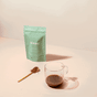 A light blue blume package stands on a surface beside a glass of minty Mint Cocoa Blend. A gold spoon with superfood powder lies nearby, set against a pale pink background.
