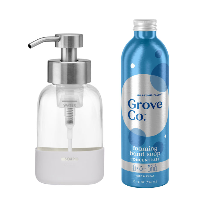 Next to a clear glass soap dispenser with a metal pump and white base is a 12 fl oz blue bottle labeled Grove Co. Foaming Hand Soap Starter Set. The design and text highlight the commitment to reducing plastic waste, reflecting Grove Co.s dedication to sustainability.