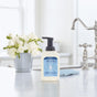 A bottle of Mrs. Meyer’s Clean Day foaming hand soap sits on a white kitchen countertop, with a silver faucet, a blue cloth, and a vase of white flowers in the background.