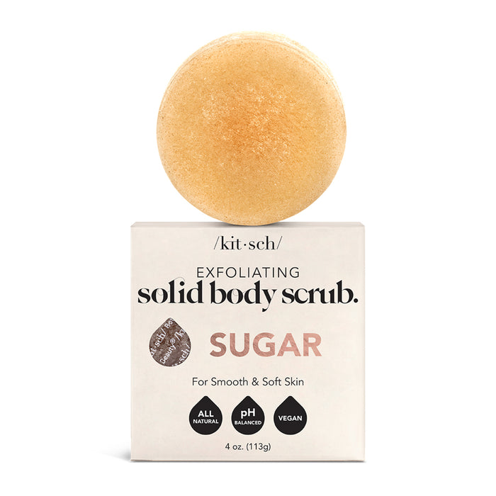 A round, beige solid body scrub sits on top of a white box labeled “/kit·sch/ exfoliating solid body scrub. SUGAR. For smooth & soft skin. The packaging highlights that it is all natural, pH balanced, and vegan.