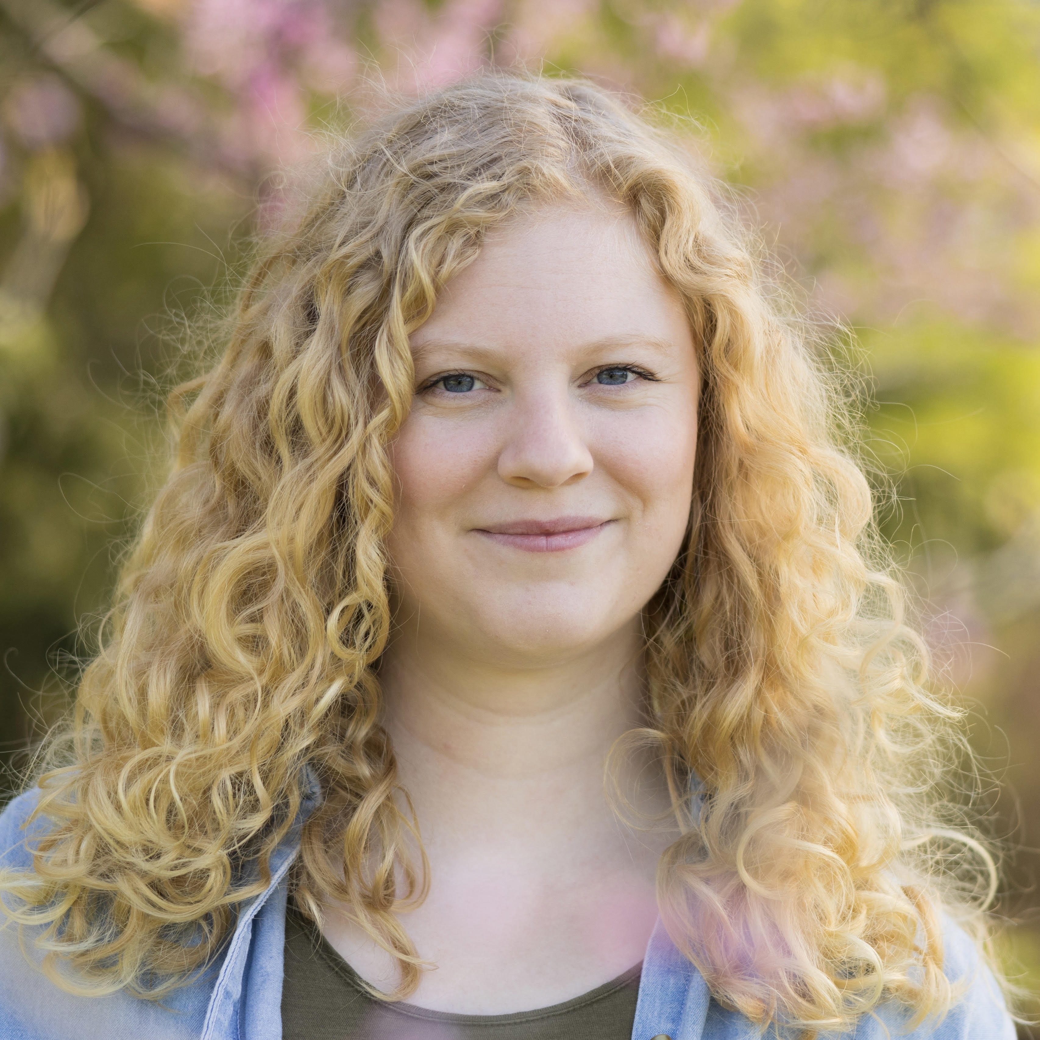 A woman with long, curly blonde hair smiles softly at the camera outdoors, with blurred green and pink foliage in the background. She wears a green top and a light blue shirt.