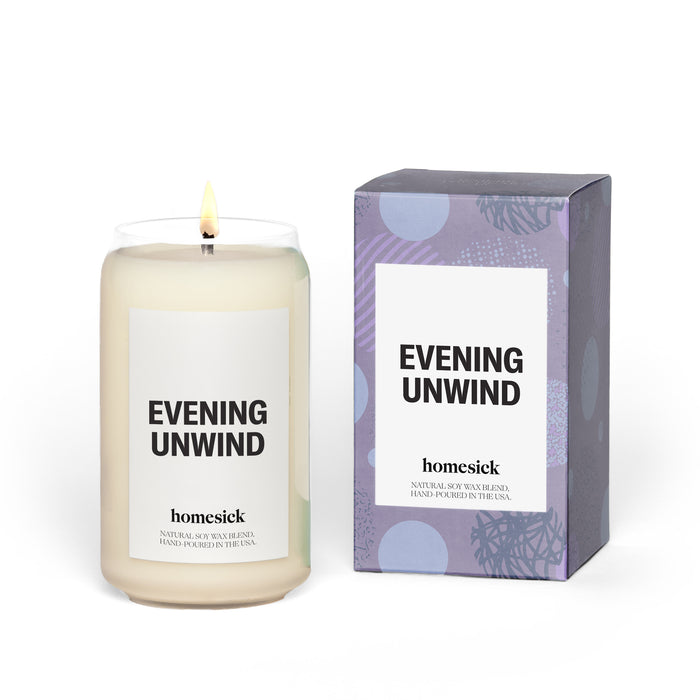 A lit white candle in a glass jar labeled Evening Unwind sits beside its purple and blue patterned box with the same label. The brand homesick is printed below the candles label.