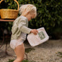 A toddler wearing a diaper and a light green cardigan is holding a pack of diapers while walking outdoors on a gravel surface. A wicker basket and green foliage are in the background.
