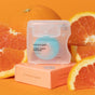 A container of Cocofloss dental floss in Cara Cara Orange flavor sits on a peach-colored box, surrounded by sliced oranges, against an orange background.