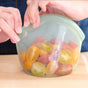 Hands close a Food Huggers Hugger Bag filled with vibrant red, yellow, and green cherry tomato halves on a wooden surface. The reusable silicone bag is easy to clean and dishwasher safe, ideal for everyday use.