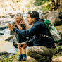 A man helps a young child drink from a water bottle while kneeling by a river in a wooded area. Both are wearing backpacks and outdoor clothing, enjoying a nature hike together.