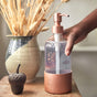 A hand pumps soap from a clear dispenser with a copper base and top, placed beside a small acorn ornament and a vase of dried wheat stalks on a wooden surface.