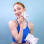 A red-haired person in a ponytail, wearing a blue tank top, smiles as they use an organic Natracare Cleansing Make Up Removal Wipe on their cheek, holding the COSMOS-certified packet against a light blue background.