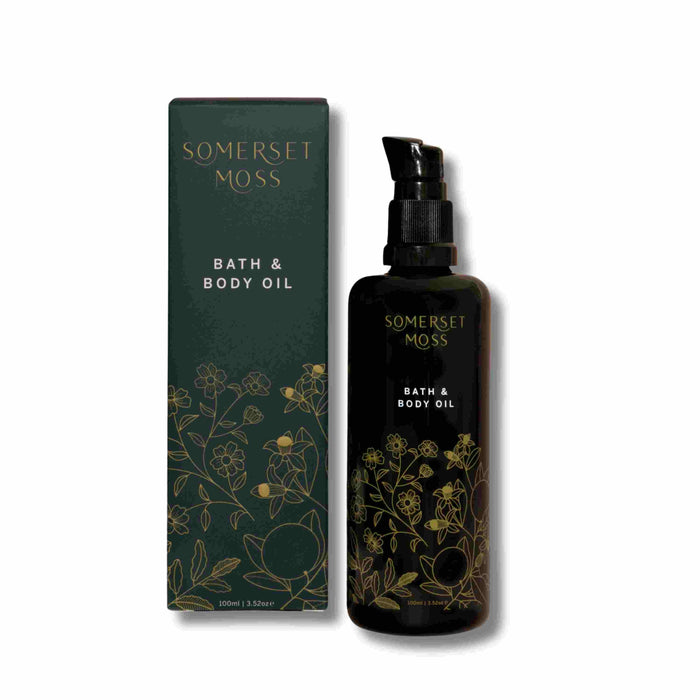 A bottle of Somerset Moss Bath & Body Oil with a pump dispenser stands next to its dark green box, both decorated with gold floral illustrations. The text on each reads Somerset Moss Bath & Body Oil.