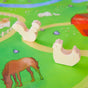 The Play Map by Sarahs Silks features wooden rooster and hen figures on a colorful farm-themed playmat with illustrations of a grazing horse and red barn. The vibrant scene transforms into a playsilk landscape, inspiring endless imaginative play.