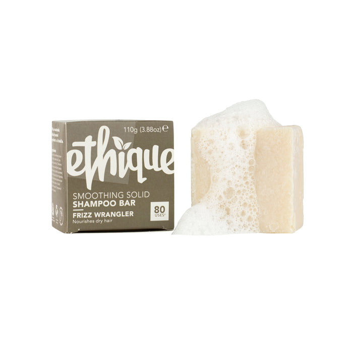 A beige Ethique Frizz Wrangler smoothing solid shampoo bar with soap suds sits beside its brown packaging, which displays product details and claims to nourish dry hair with 80 uses per bar.