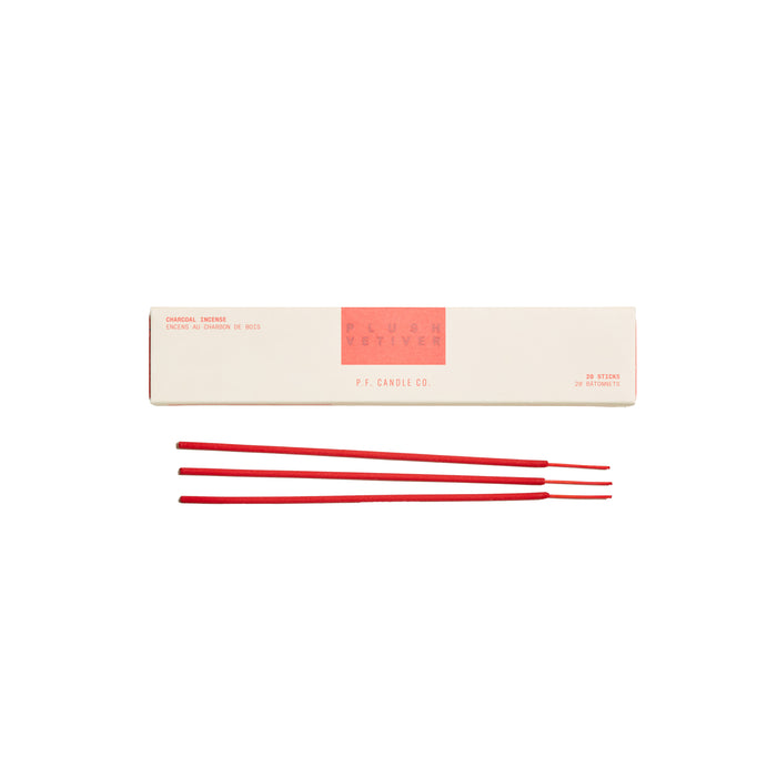 A rectangular white box labeled Plum + Vetiver P.F. Candle Co. with three red incense sticks placed in front of it on a white background.