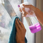 A person cleans a glass window with a blue cloth in one hand and sprays cleaner from a clear bottle with a pink base using the other hand. Sunlight shines through the window.