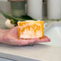 A hand holds a Soapymania Mango Coconut Soap Bar with yellow and white swirls, covered in bubbles, over a sink. In the background are blurred bathroom items like a green container and a leafy plant.