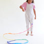 A child in a pink shirt and white overalls twirls Sarahs Silks Rainbow Streamer, creating vibrant swirl patterns on the floor against a plain white background, sparking imaginative play.