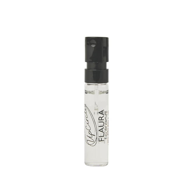 A small clear spray vial with a black cap, containing a liquid labeled Flaura Eau de Parfum and a signature-style text on the bottle.