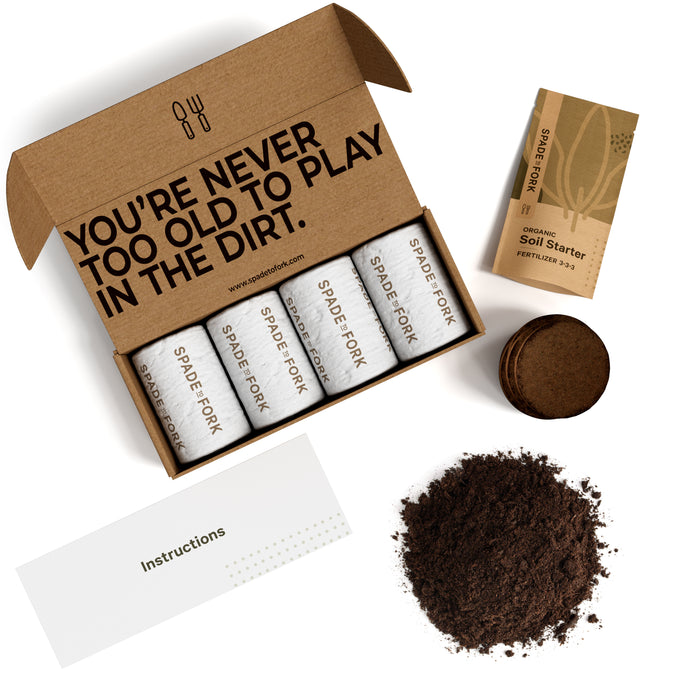 A gardening kit with four labeled seed tubes in an open box that reads YOURE NEVER TOO OLD TO PLAY IN THE DIRT, a soil starter pack, round soil discs, a pile of soil, and an instructions card.