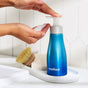 A person dispenses foaming soap from a blue Method bottle onto their hand, with a cleaning brush and sponge resting on a white tray in the background.