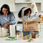 In a kitchen, a woman and child organize eco-friendly paper products. The child happily holds a roll of Bamboo Paper Towels (140 sheets per roll), while various Grove Co. boxes and packets are on the countertop. A cardboard box states, Were moving beyond plastic.