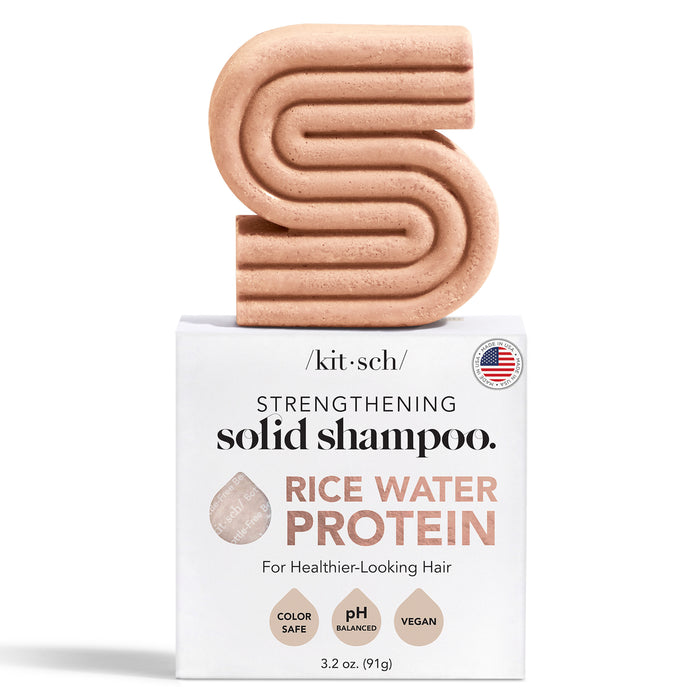 A peach-colored, S-shaped solid shampoo bar sits on top of a white box labeled Strengthening Solid Shampoo. Rice Water Protein for healthier-looking hair. The packaging highlights that it is color safe, pH balanced, and vegan.