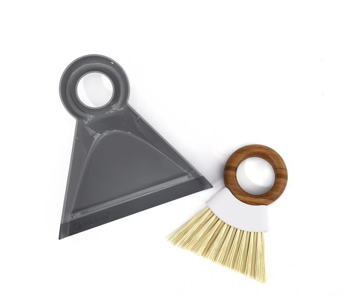 A grey dustpan with a circular handle and a small hand broom with a wooden circular handle and light bristles, both placed on a white background.