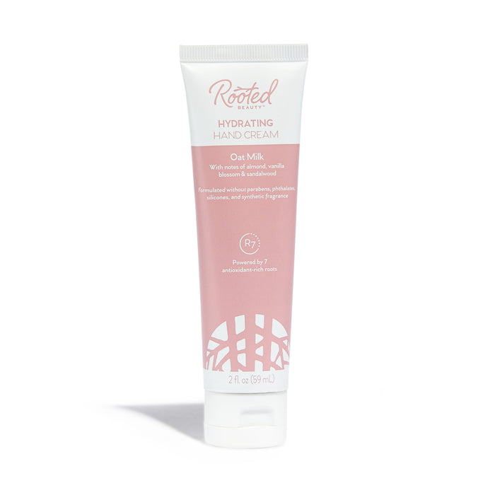 A tube of Rooted Beauty Hydrating Hand Cream in oat milk scent, featuring a pink and white design with product details printed on the front. The tube stands upright on a white background.