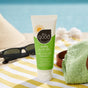 An Ultra Moisturizing Aloe Gel by All Good sits on a beach towel next to sunglasses and a green cloth, with a blurred beach and ocean backdrop creating a summery, coastal vibe.