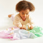 A young child with curly hair lies on a white surface, engaging with pastel Montessori toys and Sarahs Silks in pink, purple, and mint green. Reusable and fillable wooden Easter eggs made from FSC-certified wood add a natural touch. The child wears a light outfit, focused on playtime.
