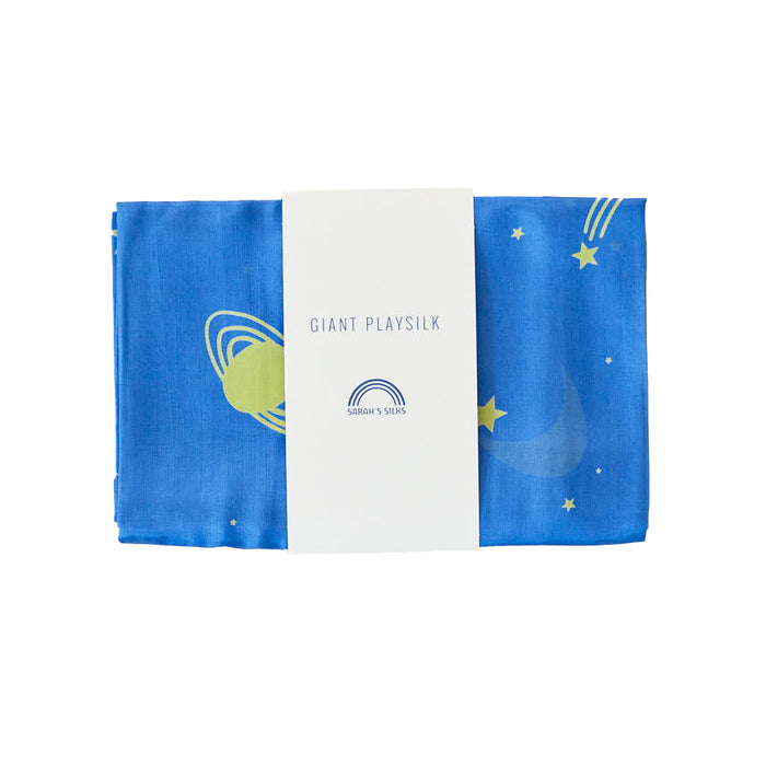 A folded space-themed blue playsilk from Sarahs Silks, adorned with stars, a crescent moon, and planets for open-ended play in Montessori & Waldorf homes. It features a white paper band labeled Giant Playsilk for Open-Ended, Imaginative Play.
