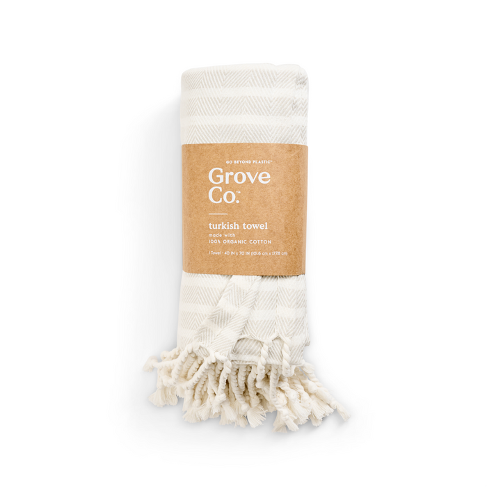 A folded ivory Turkish towel with beige stripes, labeled Grove Co. from the Spring Collection, features an organic cotton tag. It has tasseled edges and is wrapped in a brown eco-friendly paper band emphasizing its sustainable commitment.