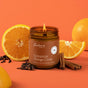 A lit Fontana Candle Co. Jar Candle – Beeswax and Coconut Oil, featuring a natural cinnamon orange clove scent, sits in front of sliced oranges, cinnamon sticks, and cloves on an orange background.