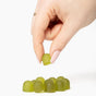 A hand with light pink nails and a gold ring picks up a Plant People WonderGreens Kids Veggie Gummy from a group of green gummies on a white background.